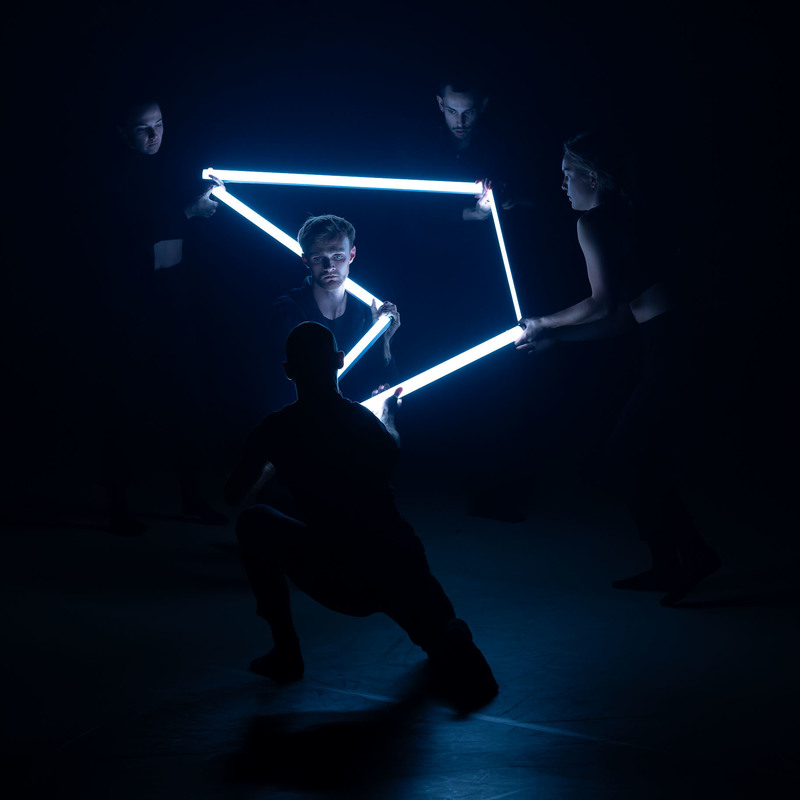 4 dancers stand in position in a textured projection and lighting state with 4 tubes of fluorescent light