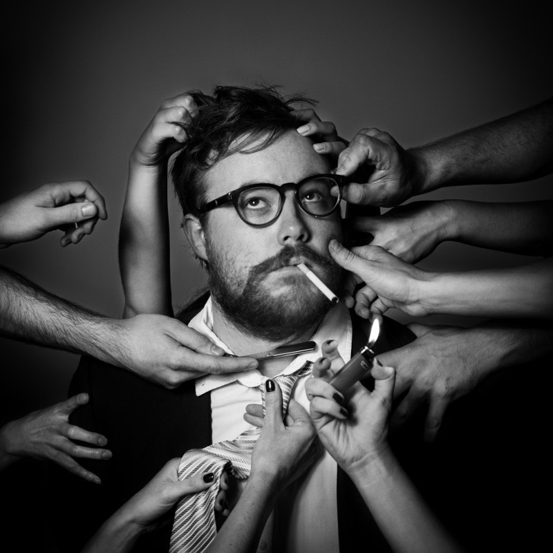 Black and white photo of comedian James Donald Forbes McCann, with several people's hands reaching into frame, one adjusting his glasses, another holding a straight razor to his throat, and another lighting a cigarette he has in his mouth.