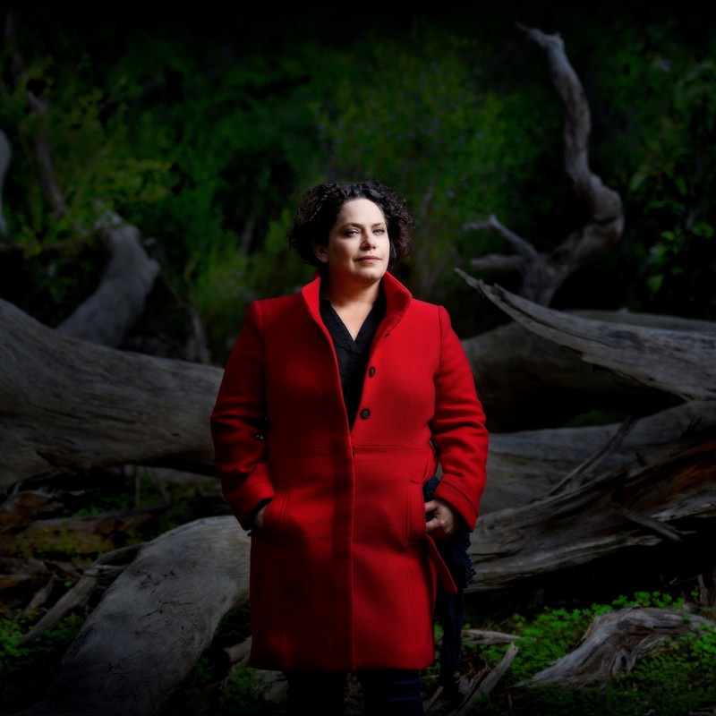 Performer Nancy Bates stands before a fallen tree in the Belair National Park with soft afternoon sunlight shining.  She stands alone, in centre shot, dressed in a bright red jacket, blue jeans, revealing a small smile, green eyes, and curly short dark brown hair.