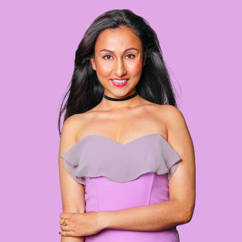 Urvi Went To An All Girls School - Urvi is of Indian heritage with brown eyes and black hair. Here she is in a pink strapless dress against a purple background. She is looking at the camera  with a smile on her face, her left hand holding her right elbow.