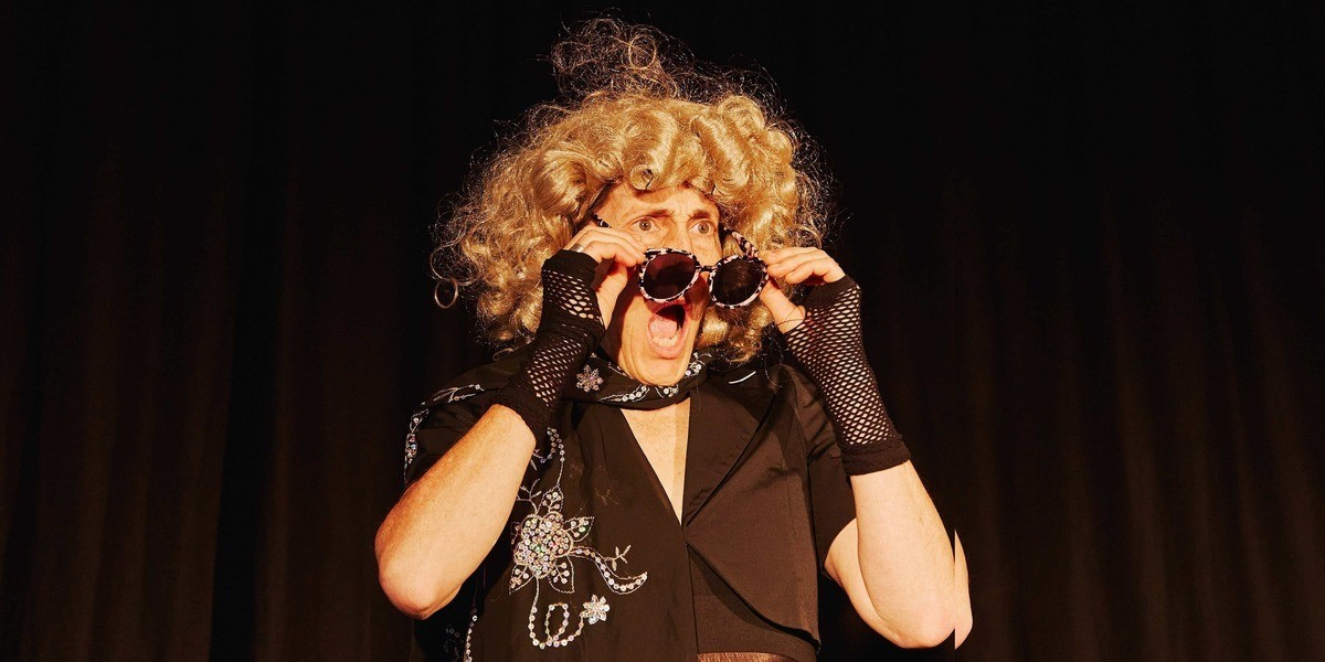 A woman in a dark dress, black lace gloves and scarf, and shining gold wig removes her sunglasses as she stares in shock, her mouth open.