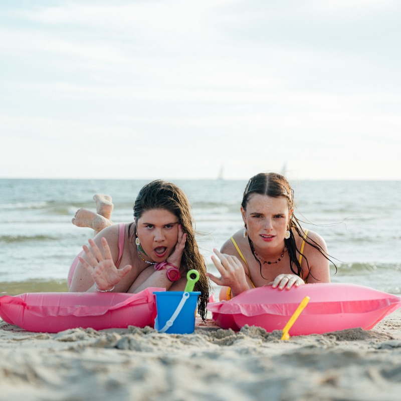 Two women lie on their stomachs on hot pink inflatable lilos on the beach. They have shocked expressions on their faces and gesture as if they are gossiping. They have wet hair and are wearing pink and yellow bikinis.