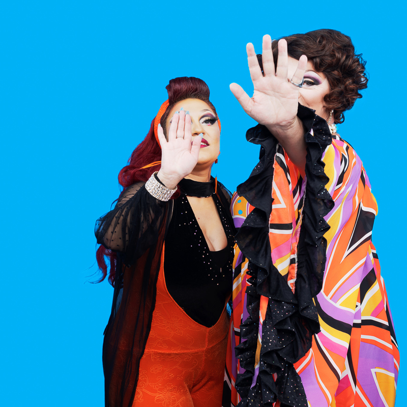 Two queens stand in front of a blue background with their hands over their faces as if to block cameras.