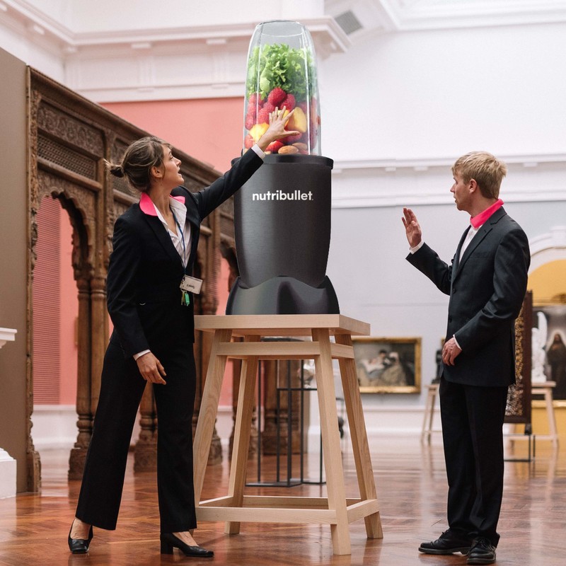 A woman and man dressed in suits gesture at a blender sitting on a plinth in an art gallery