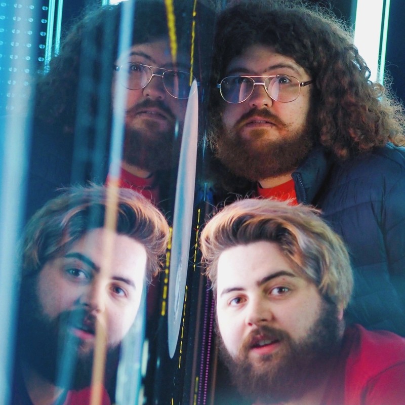 A photo of two men leaning on a wall. The man at the front has medium length hair that has been pushed over to one side and a full beard. The man at the back has long curly hair and a full beard and is wearing rose gold framed glasses. Their reflections are visible on the wall.