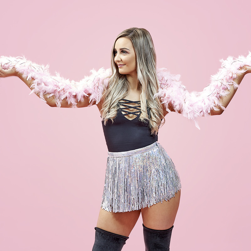 Less Bitter, More Glitter - Jayne of June stands in front of a pink background and holds up a pink feather boa in both hands while wearing a black top, glitter skirt and black knee high boots.