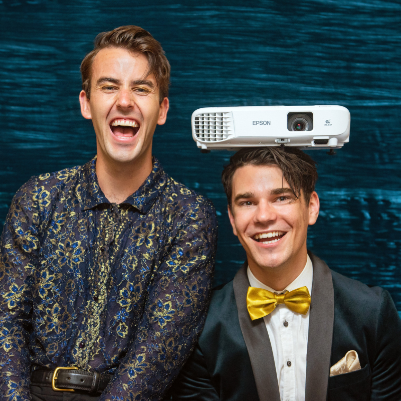 Two well-dressed men sit side by side, smiling. One has a projector sitting on his head.
