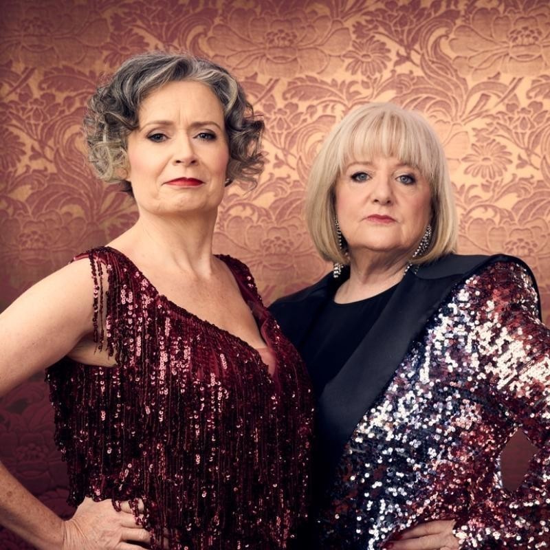 JUDITH LUCY & DENISE SCOTT - STILL HERE - Mid shot of Judith Lucy and Denise Scott staring defiantly at the camera. Both have hands on hips. Judith is wearing a sequined burgundy sleeveless dress and Denise is wearing a black and red sequined jacket with dangly sparkly earrings. They are standing in front of a gold and red flocked wallpaper with and antique floral pattern.