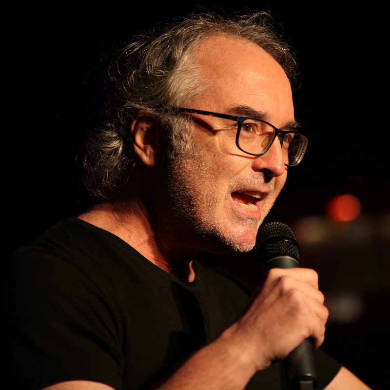 Justin has short silver hair and facial stubble. He is wearing black framed reading glasses and a black T-shirt. Justin is facing away from the camera and speaking into a black microphone.