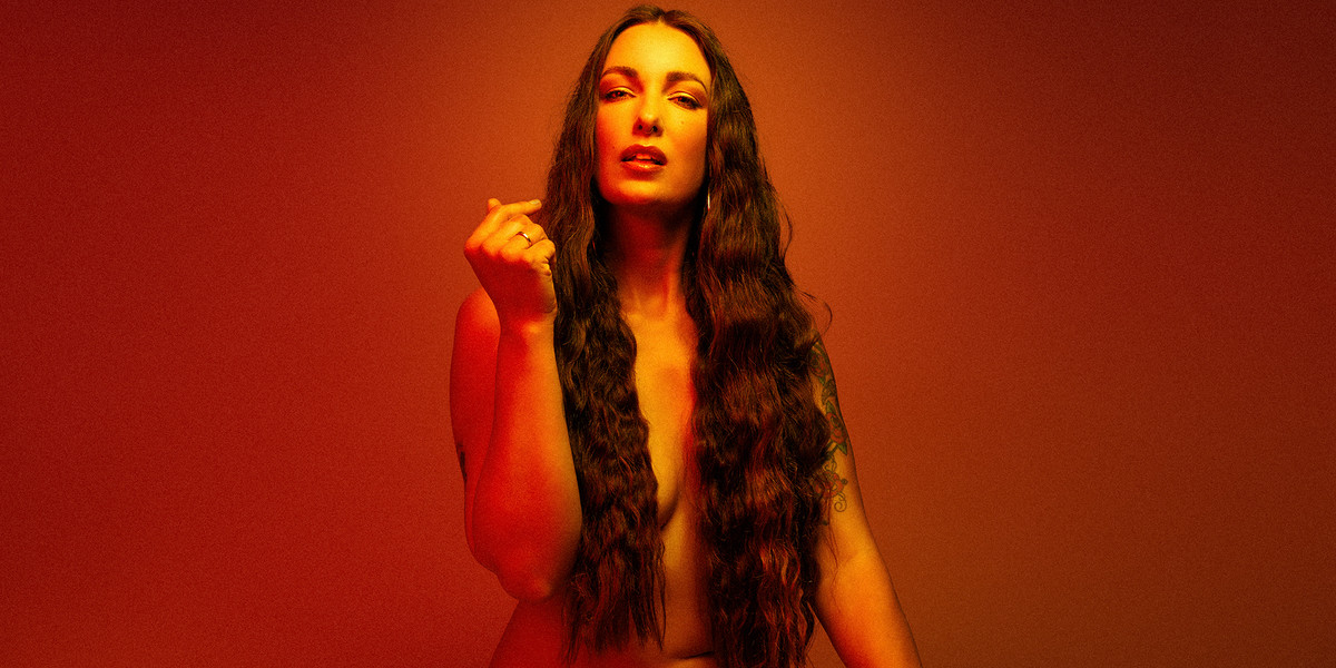 A Body At Work - A cis gendered female squats down on seven-inch lucite heels wearing no other clothing. She has long dark hair which completely covers her breasts and is gazing straight at the camera. She is bathed in warm orange and golden light and sits against s deep burnt orange and golden yellow backdrop.