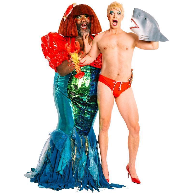 Le Gateau Chocolat who is a 6 foot tall, plus sized drag queen with Black skin is dressed as a mermaid. Wearing a large red wig, bright rosy makeup, a red vinyl bikini, and green& blue sequin mermaid dress, while looking adoringly to the right with his hand reaching gently out. To his right, Jonny Woo, who is a 6 foot drag queen with white skin and a slim muscled body, wears a blonde choppy wig, electric blue eyeshadow, and only a small red speedo and matching red stiletto high heels. Jonny has a shocked look on his face as he is sandwiched between the mermaid and a grey rubber shark face which Jonny holds on his left hand like a puppet.