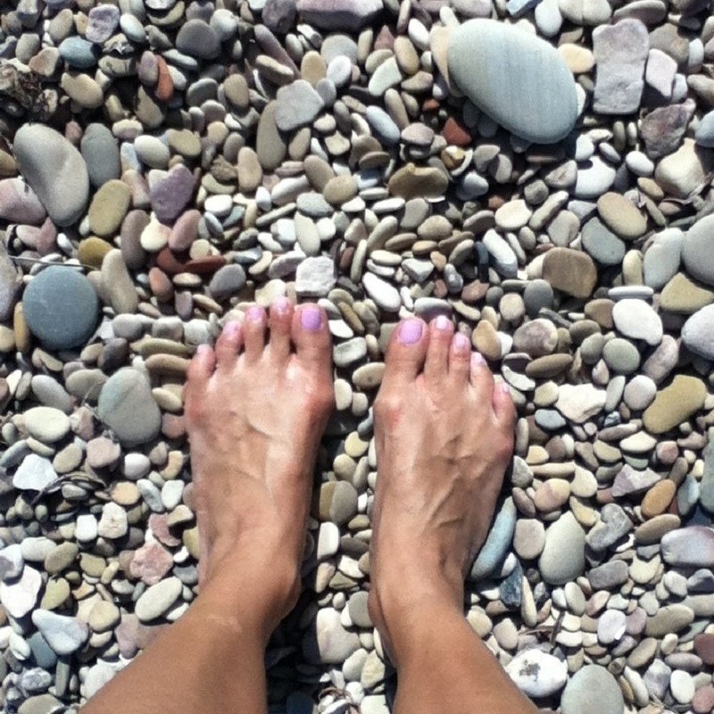 Song Revisited - A photo of a person’s feet standing on pebbles of various sizes shapes and colours. They have pink nail polish on their nails.