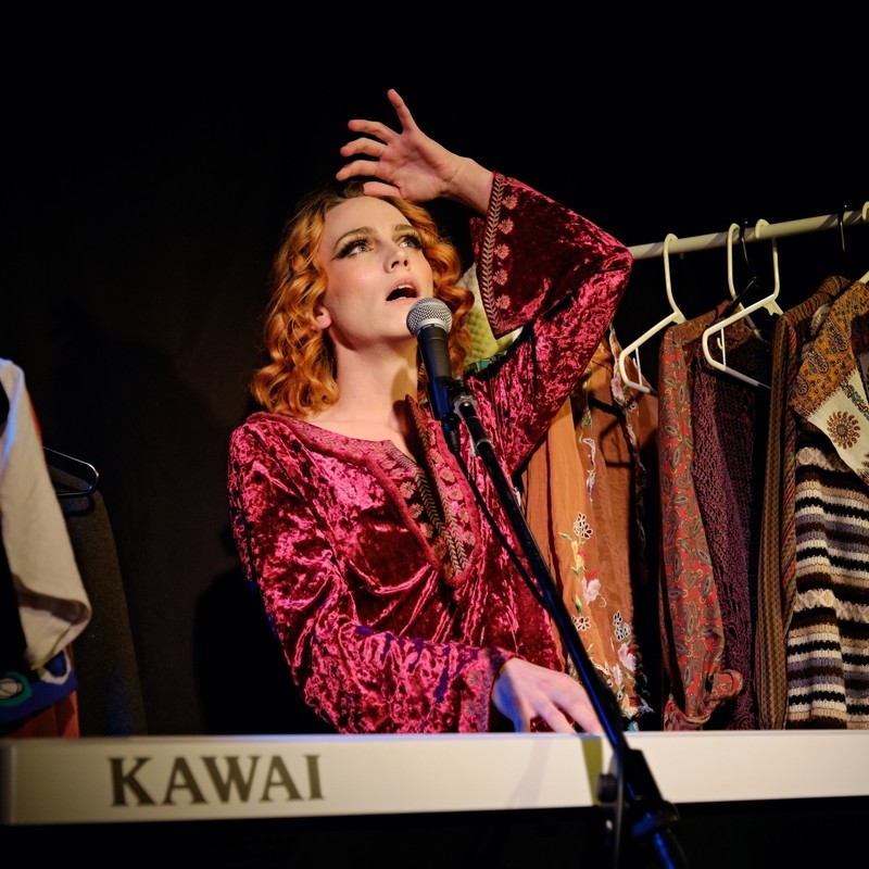 The Alchemist of Feelings - Frankly is pictured performing live. She is looking upwards while singing into a microphone. One of her hands is stretching above her head. She is seen with curly, orange hair, wearing a pink velvet blouse and heavy eye make up. A rack of clothing can be seen behind her.
