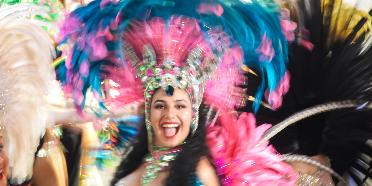 BARRIO BEATS Latin Fest - Black haired carnival Samba dancer in pink and blue feathered headdress, smiling in glee