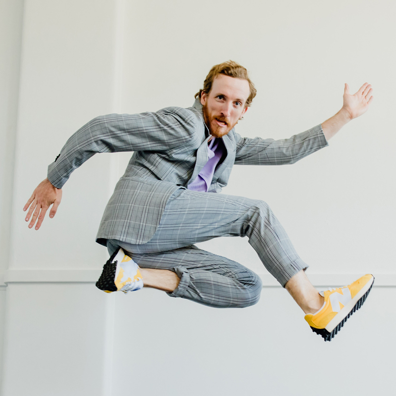 Will Tredinnick, the 1-man performer of Pickled Sink, jumps into the air while looking at the viewer. He is wearing a grey suit and bright yellow sneakers.