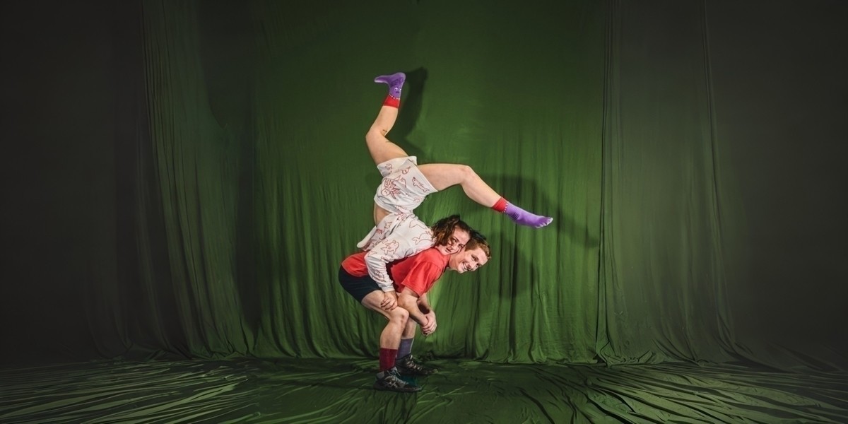 A performer balances on the back of another. There is a green curtain behind them.