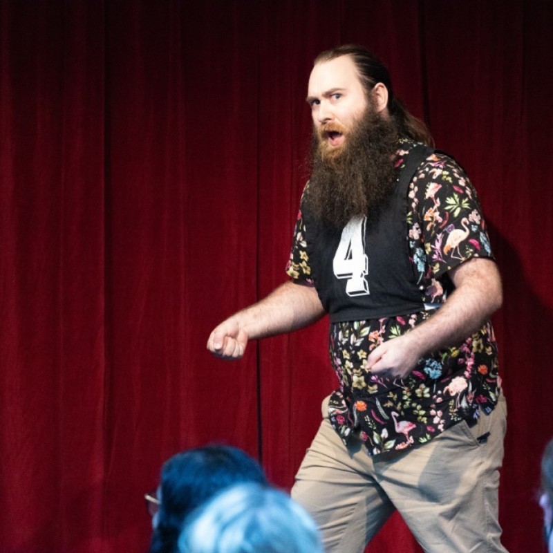 A bearded performer is in mid-song and miming trimming a hedge on stage.