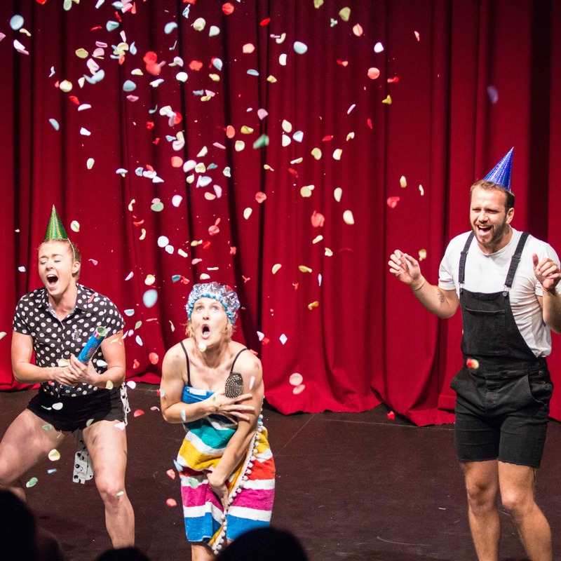Three people stand in front of a red curtain backdrop. The person in the middle is dressed in a rainbow towel and shower cap. The other two have party hats on their heads. All are screaming and confetti is falling from the ceiling.