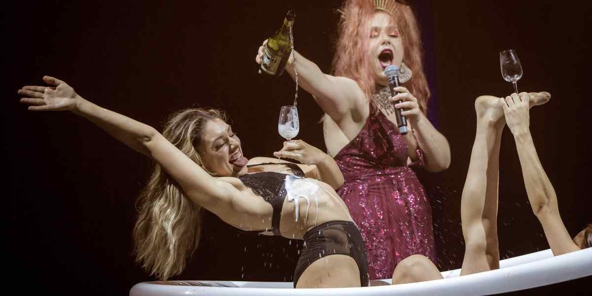 Dancers are sitting, laughing in clawfoot bathtub as Eliza (singer) pours champagne over dancers whilst singing