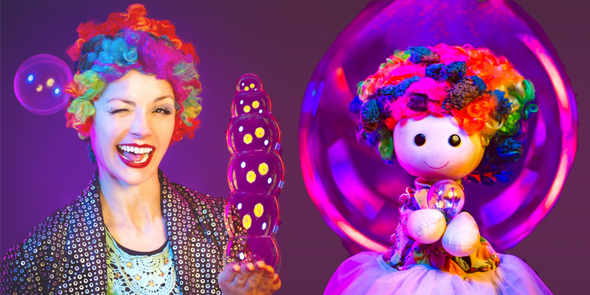 A female clown stands next to a puppet that looks like a small version of her, the female clown holds a stack of floating bubbles the tiny puppet holds a single bubble with a large bubble floating behind her.