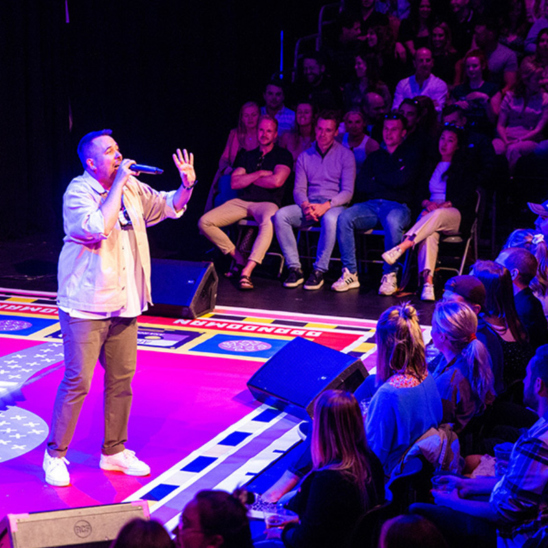 Rob is standing on a brightly lit stage, talking to an audience member higher in the audience rake. We can see the audience on one side and in front of him. The stage is covered with a brightly coloured rug.