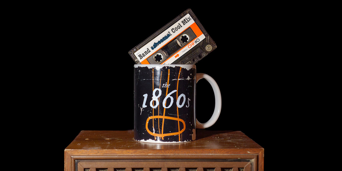 1860s: Now on Cassingle - A coffee mug on top of a wooden speaker cabinet. The coffee mug has text that says 'The 1860s' in a stylised font. A cassette tape is balanced in the coffee mug, with type on the label that says 'Band Cool Mix'.