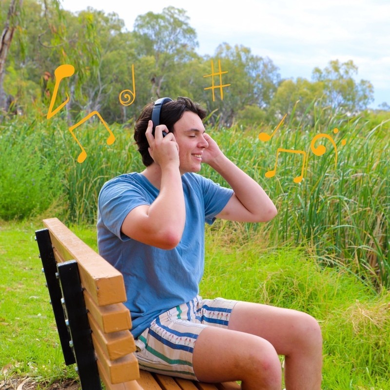 Riverland Silent Disco - A young man sits on a park bench surrounded by river reeds. He listens to music through his headphones and smiles.