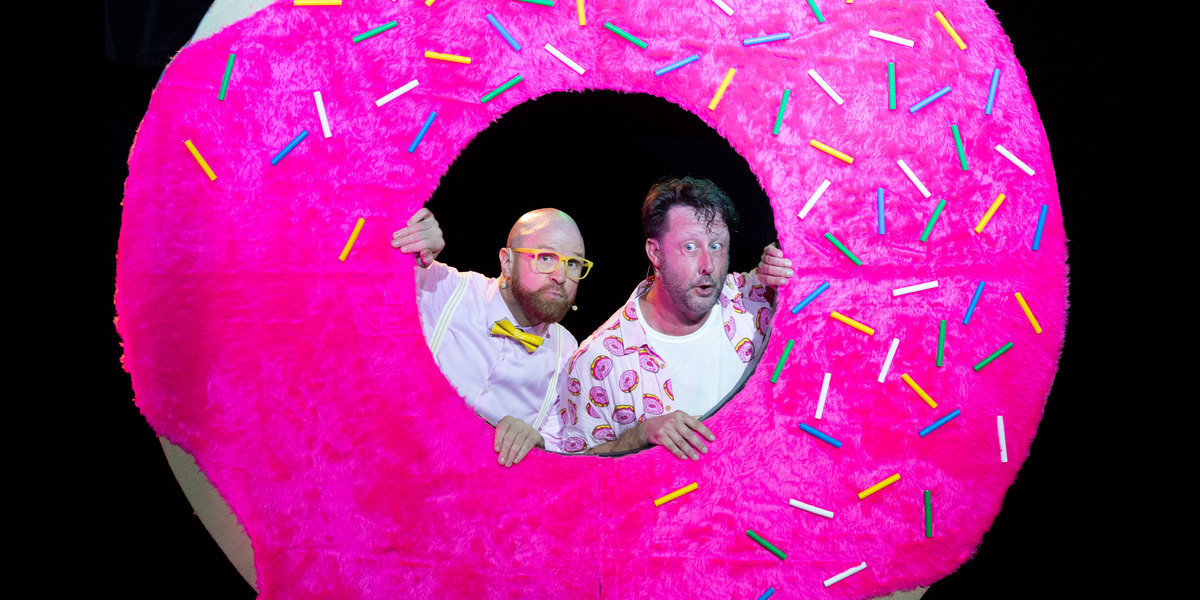 Two men in pink shirts look through the hole in a very large pink fur doughnut.