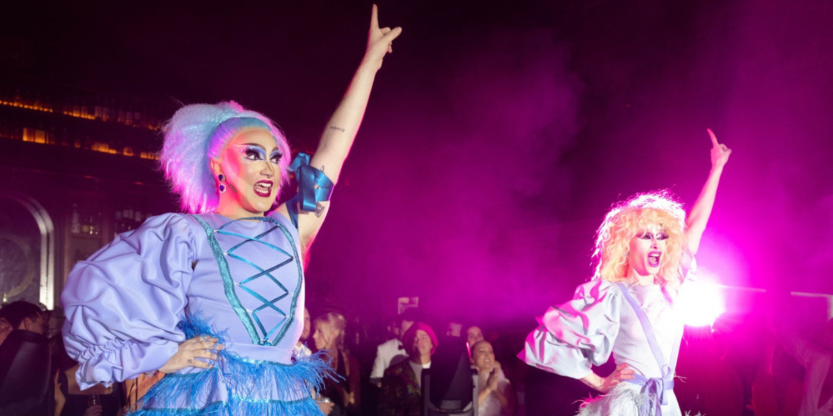 Two Drag Queens on Stage, with one arm in the arm pointing and the other on their hip, both are wearing a leotard and a skirt, one in dark blue and one in light blue. One is in a blonde wig and the other in a blue wig