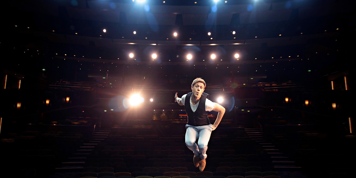 Magician Martin Brock leaps energetically into view, making dynamic gestures toward the theater behind him, while a cascade of radiant lights converges directly on the camera, all captured in his engaging gaze.