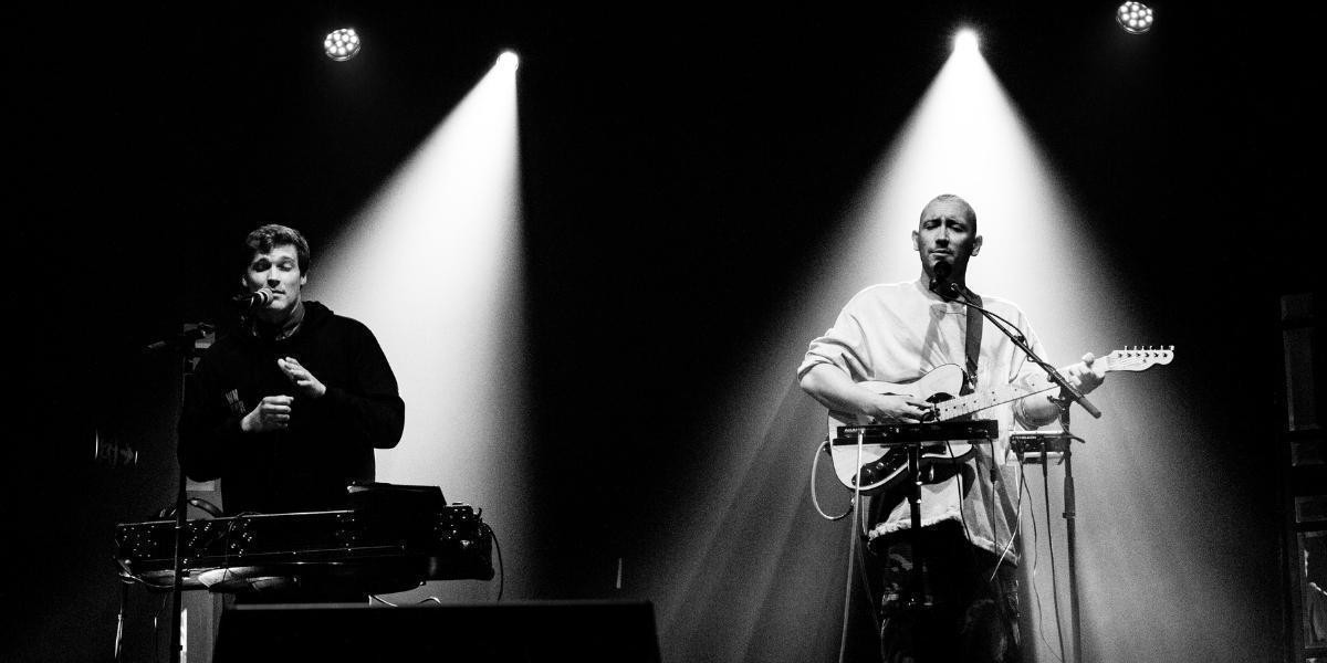 Jamie MacDowell and Tom Thum stand on a stage side by side under two individual spotlights. Portrayed in black and white, the duo are both singing with a lot of emotion. Tom is standing behind a table with looping and sampling equipment on it, Jamie is playing an electric guitar.