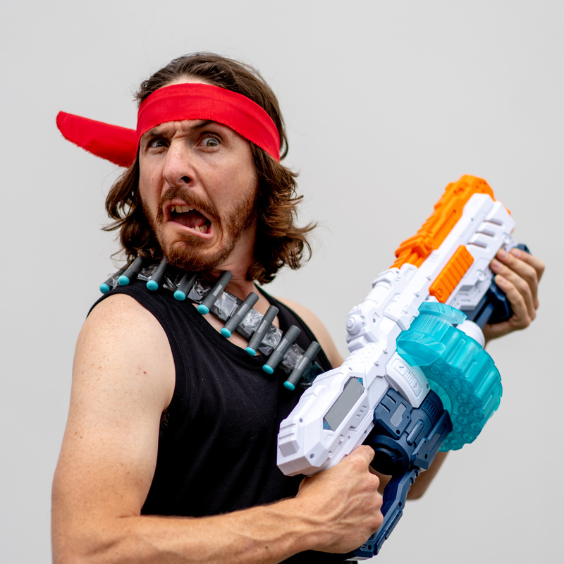 MAN-BO - A photo of a person with a bewildered expression on their face. They are wearing a black singlet and a red bandana across their forehead. They are holding a large plastic gun in front of their body.