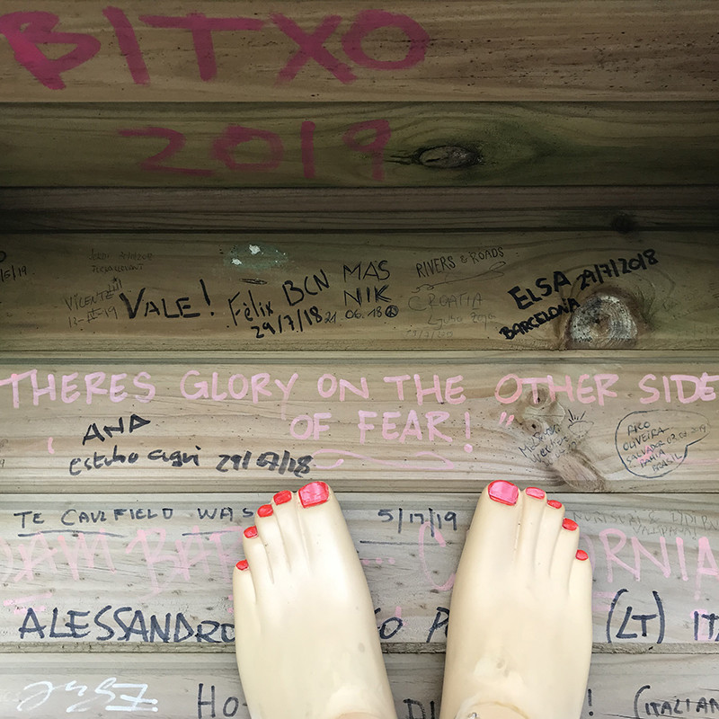 A photo of a pair of plastic feet with red nail polish on a wooden background that has lots of words written across it.