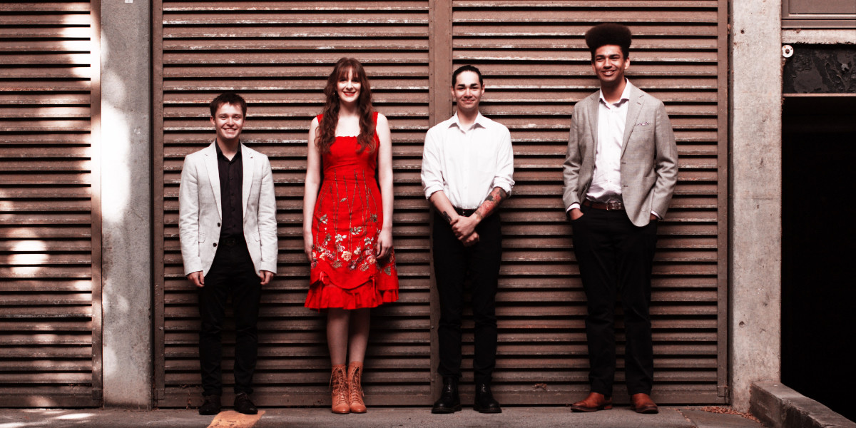 Four performers (Left to Right: Solomon Young, Tiffany Gaze, James Ho and Jackson Mack) stand in front of shuttered doors. Tiffany wears a red dress.