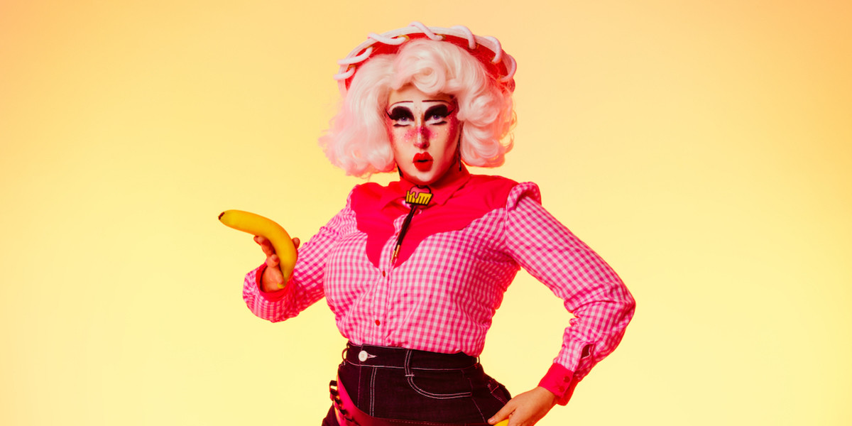 The drag queen Clara Cupcakes is pouting at the camera and holing a banana like a gun while standing in front of a sunset gradient background. She is dressed as a cowgirl in a red hat with white trim, a pink checked shirt with a pink cupcake bolo tie and denim shorts. Her hair is white and she has a full face of drag make-up.