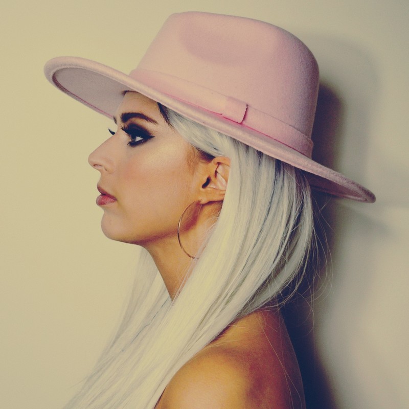 A side profile headshot of a person with platinum blonde hair wearing a pink hat.