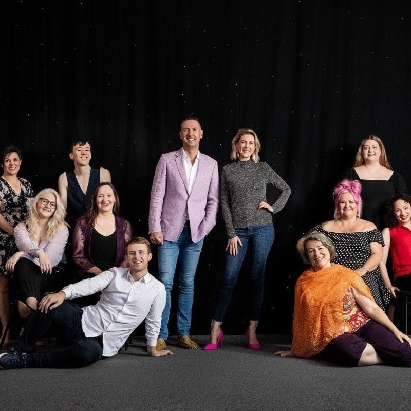 The Cabaret Lab: Lights Up! - The Cabaret Lab group facing the camera, standing in front of black curtains. In the middle are Michael Griffiths and Amelia Ryan. Michael is wearing a pink blazer, white shirt and jeans. Amelia has her left hand on her hip, wearing a gray long-sleeve shirt, jeans, and pink heels.