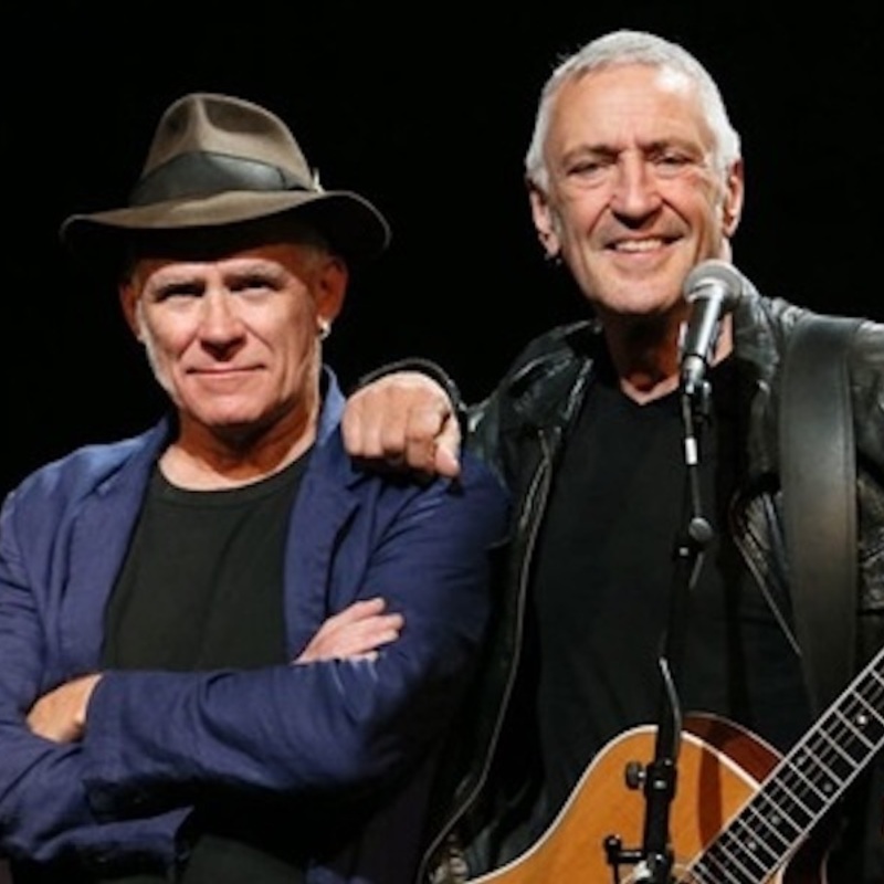 A smiling John Waters, the lead singer and star of the show, stands with a guitar slung over one side and his hand on Stewart D'Arrietta's left shoulder. Stewart is wearing a blue suit jacket over a black t-shirt and his signature hat. John is wearing a black leather jacket and black t-shirt.