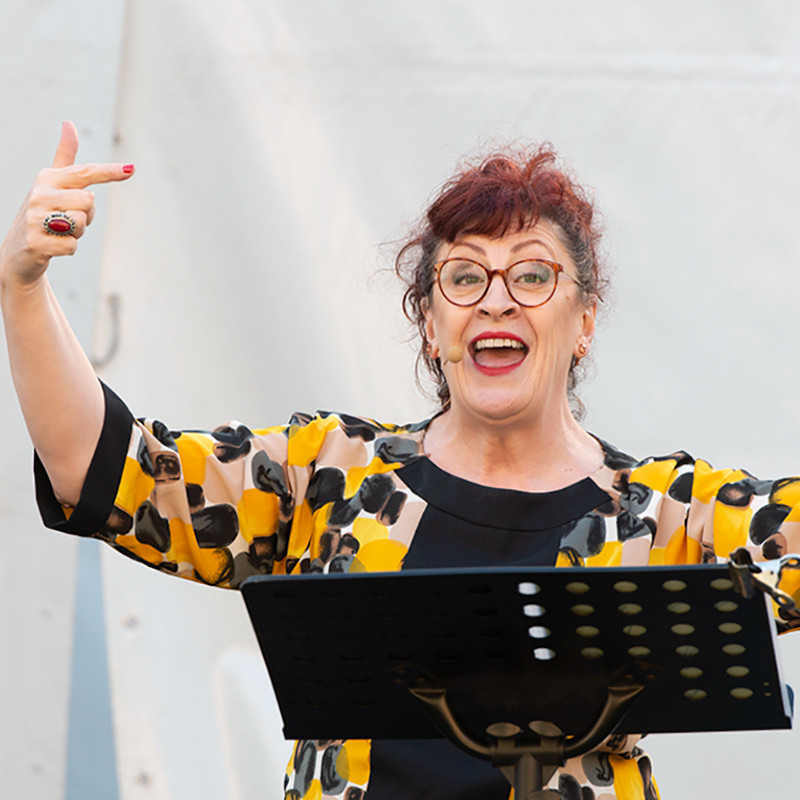 PUBsing @ The Fringe - A photo of a woman standing behind a black music stand wearing a yellow and black top and red framed glasses.