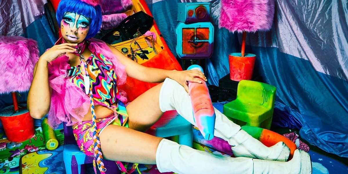 A stunning lady rave clown lounges in a psycadelic instalation of childlike objects.  the whole sceen is very colorful, as is she.