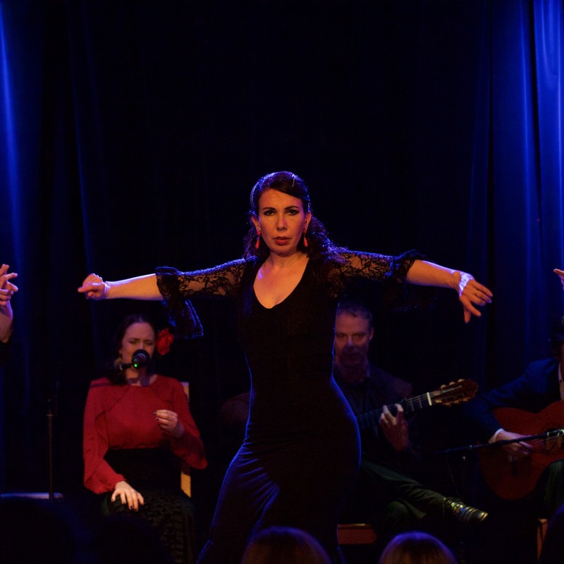 Three flamenco dancers in black costumes with musicians behind.