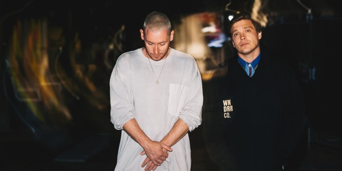 Jamie Macdowell and Tom Thum stand side by side, Jamie is wearing a longsleeve oversized white top and looking at the ground, Tom is wearing a black hoodie and looking at the viewer with a pleasant, but distant expression. There are light flares surrounding the framing of their figures.