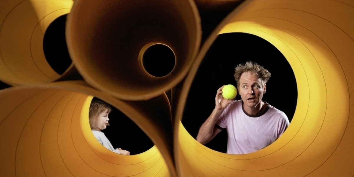 Four cardboard horizontal cylinders containing a man and a young girl. Man holding a tennis ball up to his left ear as if listening to the tennis ball. The young girl is watching the man.