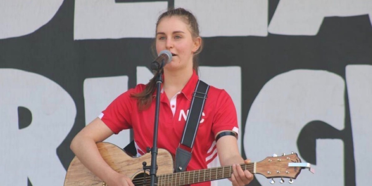 A close up image of Poppy singing. She is standing behind a microphone in the centre of the image, facing forwards. She wears a red tshirt and is holding a guitar. The background is made of black and white shapes.