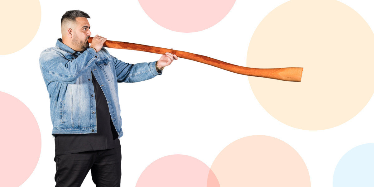 An Aboriginal man wearing a denim jacket and black jeans stands sideways playing a didgeridoo against a background of coloured circles.