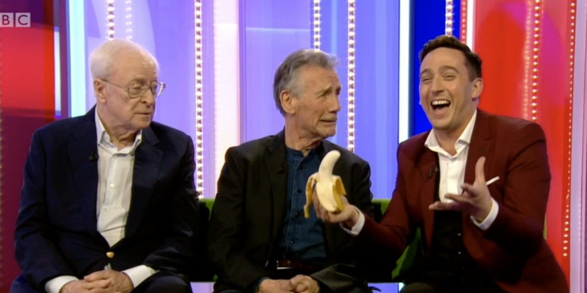 Paul is appearing on a BBC tTV show called the one show with Sir Michael Palin and Sir Michael Cane his is holding a banana as part of a magic trick and laughing while Michael Palin pulls a funny face.
