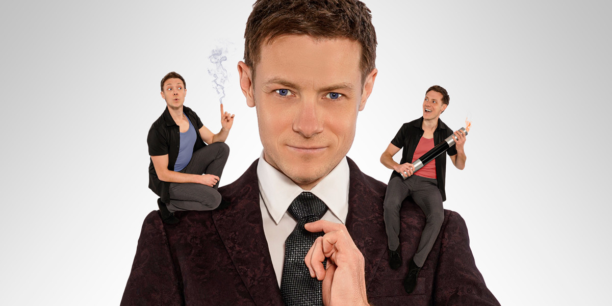 Magician Martin Brock gazes directly into the camera, accompanied by a miniature version of himself on his right shoulder, conjuring smoke from his finger, and another miniature version on his left shoulder, wielding a fiery wand with a determined expression.
