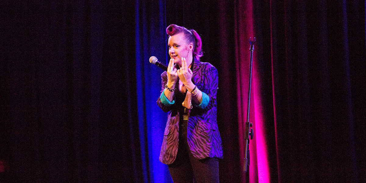 A comedian wearing a purple tiger-print blazer holds their middle and index fingers of both hands up, highlighting the deliberate difference in length.
