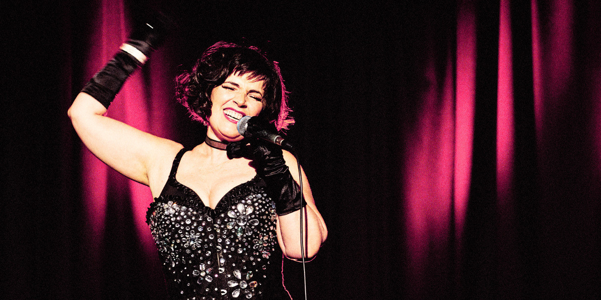 GOOD GIRL BAD GIRL - Souzi, a white woman, is looking ecstatic whilst singing on stage. She is wearing a glitzy costume comprising a black bodice heavily decorated with large silver jewels. Both hands are covered in black satin gloves to elbow, her left hand is holding a microphone near her mouth but not covering a big smile, and the right gloved hand with diamante bracelet is raised in the air. Her hair is a chin length, dark and curly bob with raggedy fringe with light shining through creating a pink halo. The background is black with pink stage lights highlighting the velvet curtains.
