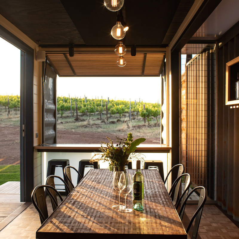 Afternoon sun is shining through a picture window, showcasing the beautiful views of the vines. Four stools are placed at the window and a long wooden table is set with wine glasses and a bottle of wine.